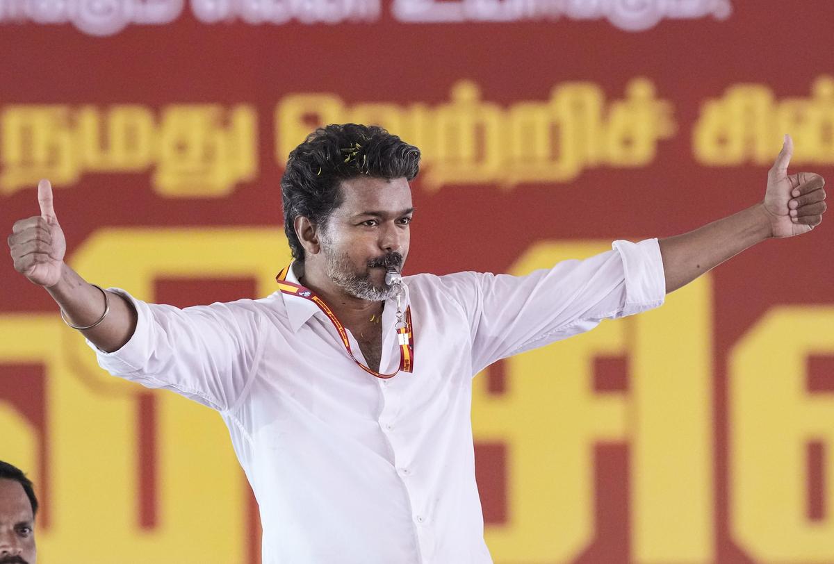 Man on stage in white shirt giving two thumbs up, wearing a lanyard, whistle in mouth, celebrating in front of a yellow-red backdrop